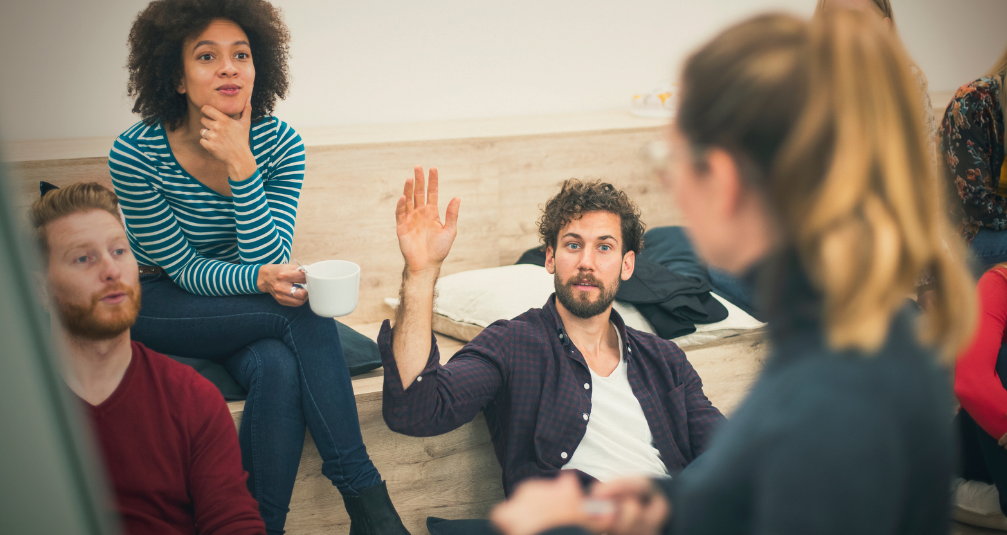 Three people look at a speaker who is standing next to a whiteboard. in the middle of the people, a man has his hand raised